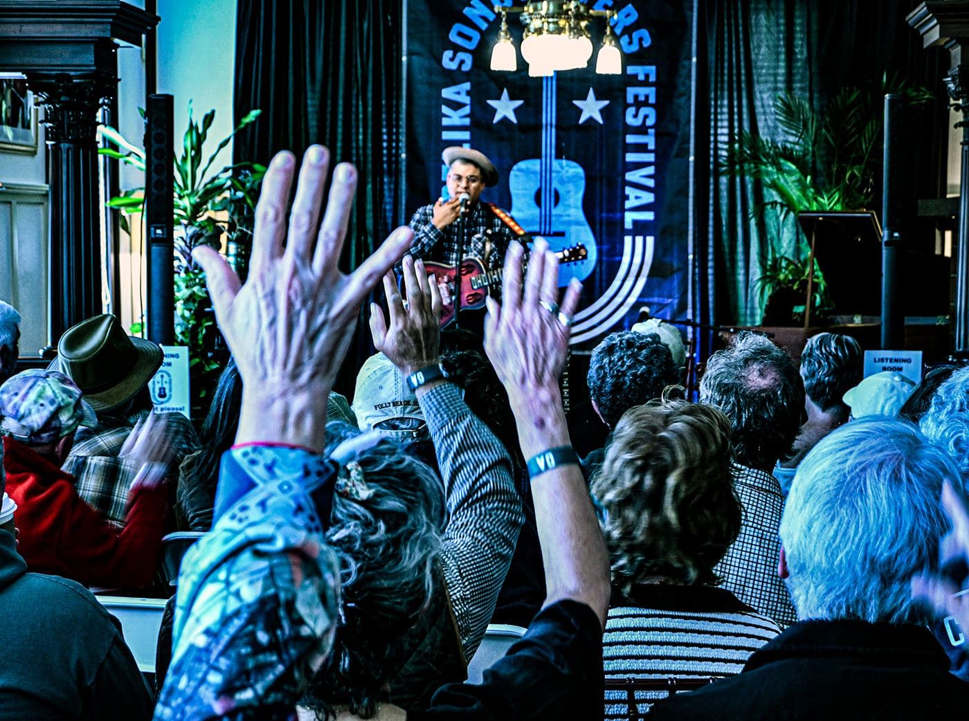 Raised hands in the audience with a songwriter on stage under the festival banner