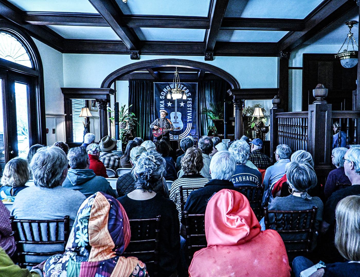 A full room of listeners in the Heritage House parlour facing the stage